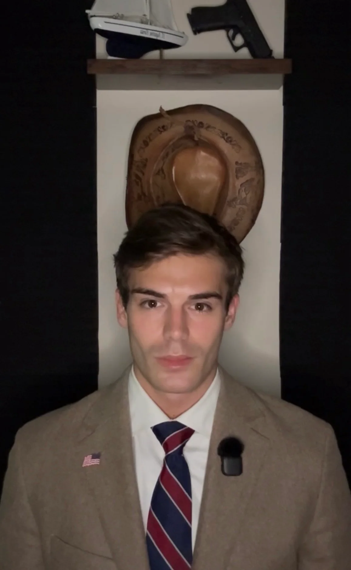 Joshua Michael in a tan blazer, white shirt and striped tie with an American flag pin, a cowboy hat on the wall behind him.